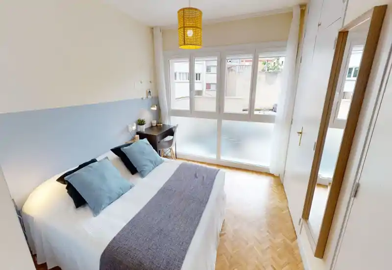 Chambre lumineuse et bien mise en scène avec un grand lit double habillé de linge blanc et coussins bleu clair. Un bureau près de la fenêtre et le parquet apportent fonctionnalité et chaleur ; de larges fenêtres offrent une belle lumière naturelle.