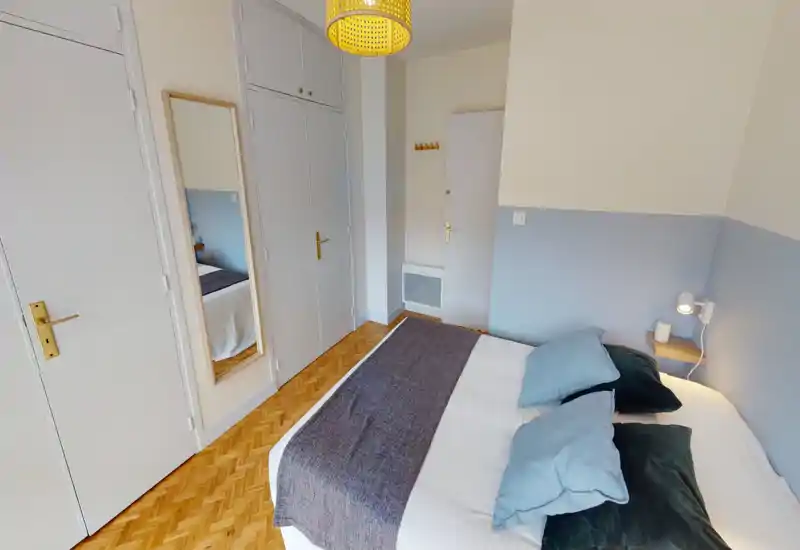 Chambre lumineuse et bien mise en scène avec un grand lit habillé de linge neutre, coussins superposés et un jeté texturé. On remarque un parquet, des placards intégrés, un miroir en pied et une petite étagère de chevet avec lampe de lecture.