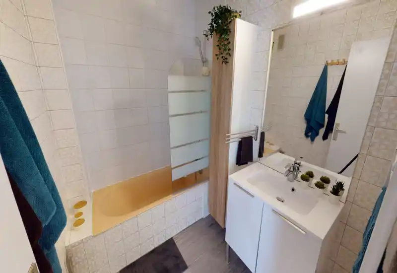Bagno pulito e ben illuminato con vasca, paratia in vetro, mobile lavabo con specchio e piccole piante decorative.