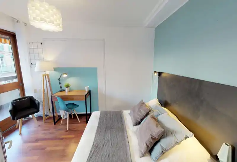 Bright, modern bedroom with a large double bed, soft cushions and coordinated teal accent wall; a small workspace with desk and chair sits by the window, plus a cozy armchair and floor lamp.