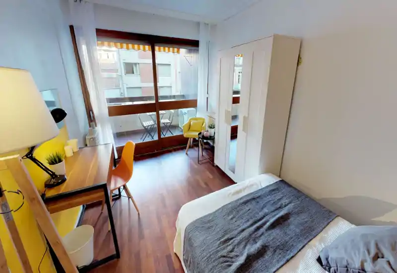 Bright, modern bedroom with a comfortable double bed, wooden floor, desk area and a balcony accessed through large glass doors — ideal for natural light and a cozy work corner.