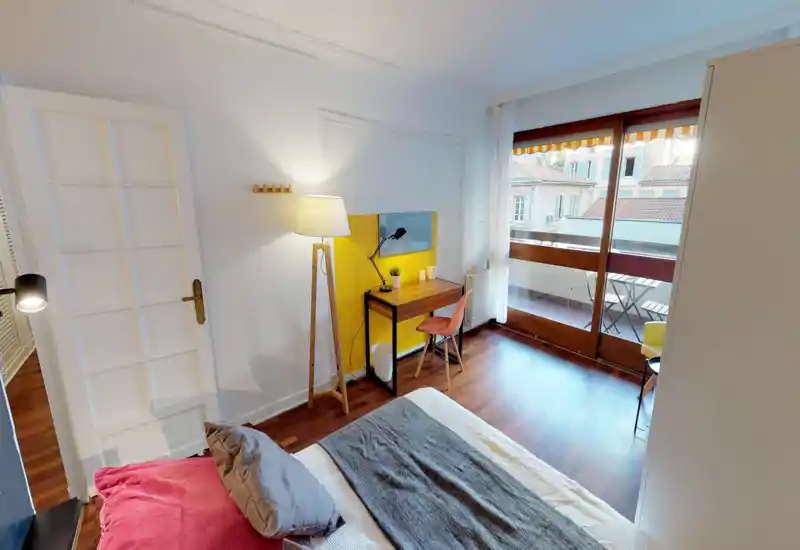 Sunny, stylish bedroom with a comfortable bed in the foreground, a wooden desk and chair beside a large balcony door that fills the room with natural light.