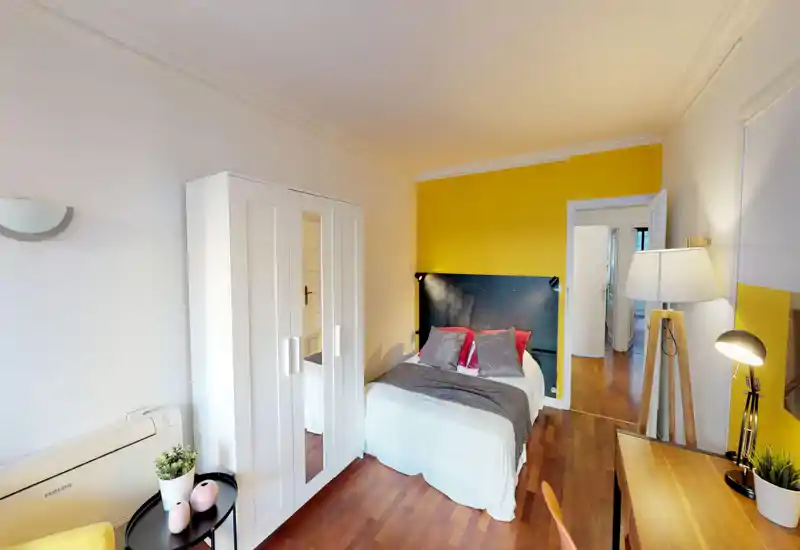 Bright, styled bedroom with a double bed, colorful cushions, wardrobe with mirrored door and a wooden floor. Warm yellow accent wall and thoughtful lighting create a cozy atmosphere.