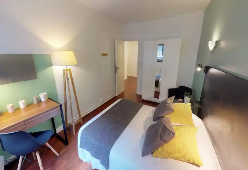 Bright, styled bedroom with a double bed dressed in grey and yellow textiles, a bedside table, reading lamp and a small desk — cozy and well-lit for rental listings.