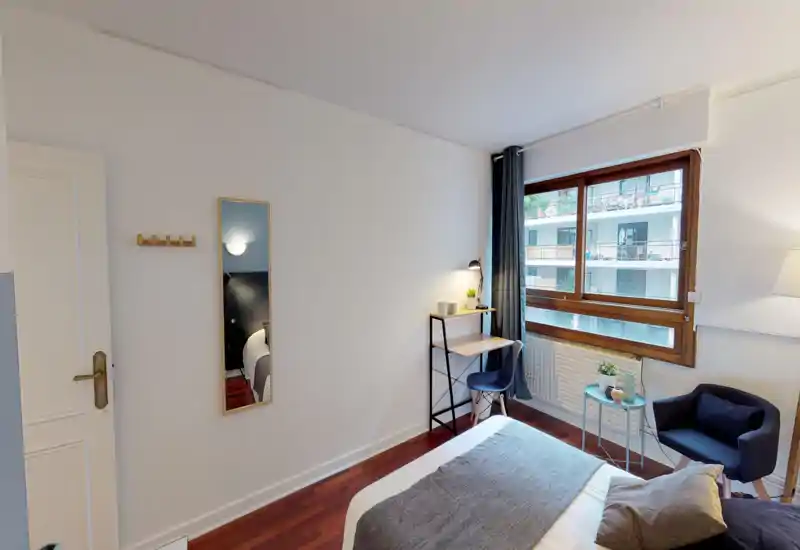 Bright, modern bedroom with a neatly made double bed, a small desk by the window, a cozy armchair and simple, stylish decor. Large window provides natural light and a pleasant street view.