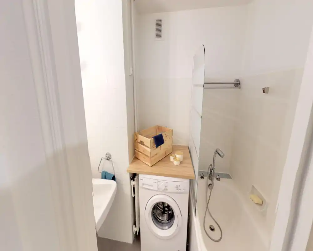Compact bathroom with a bathtub, handheld shower, sink and a washing machine tucked under a wooden countertop; neutral tiles and soft lighting create a clean, functional space.