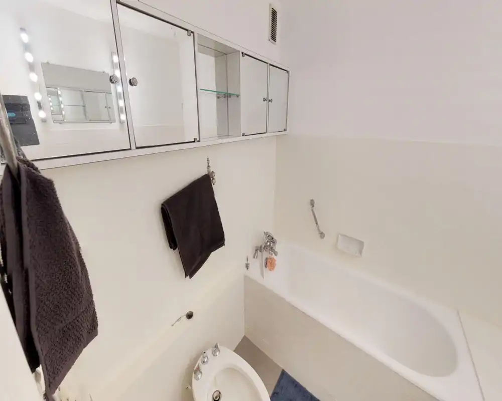 Bright, clean bathroom with a built-in bathtub, bidet and mirrored medicine cabinets above the sink area. White tiles and neutral towels give a fresh, minimalist look.