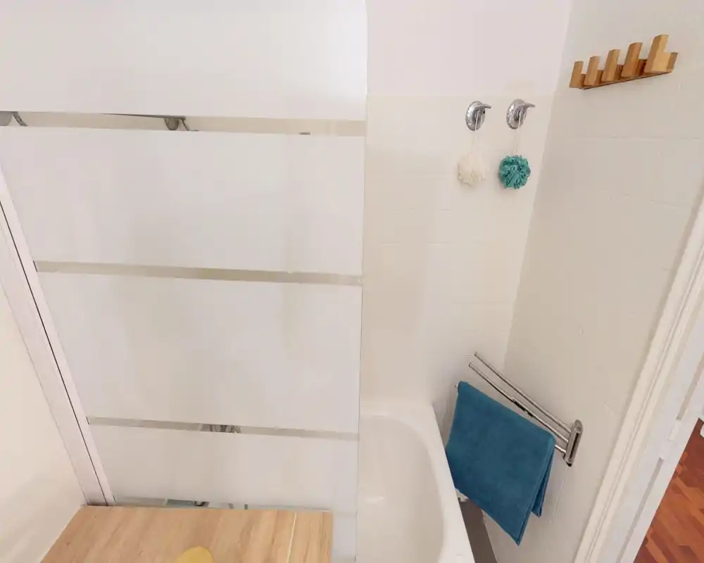 Clean, bright bathroom with a bathtub and frosted glass shower screen, wall-mounted hooks and a towel rack with a teal towel.