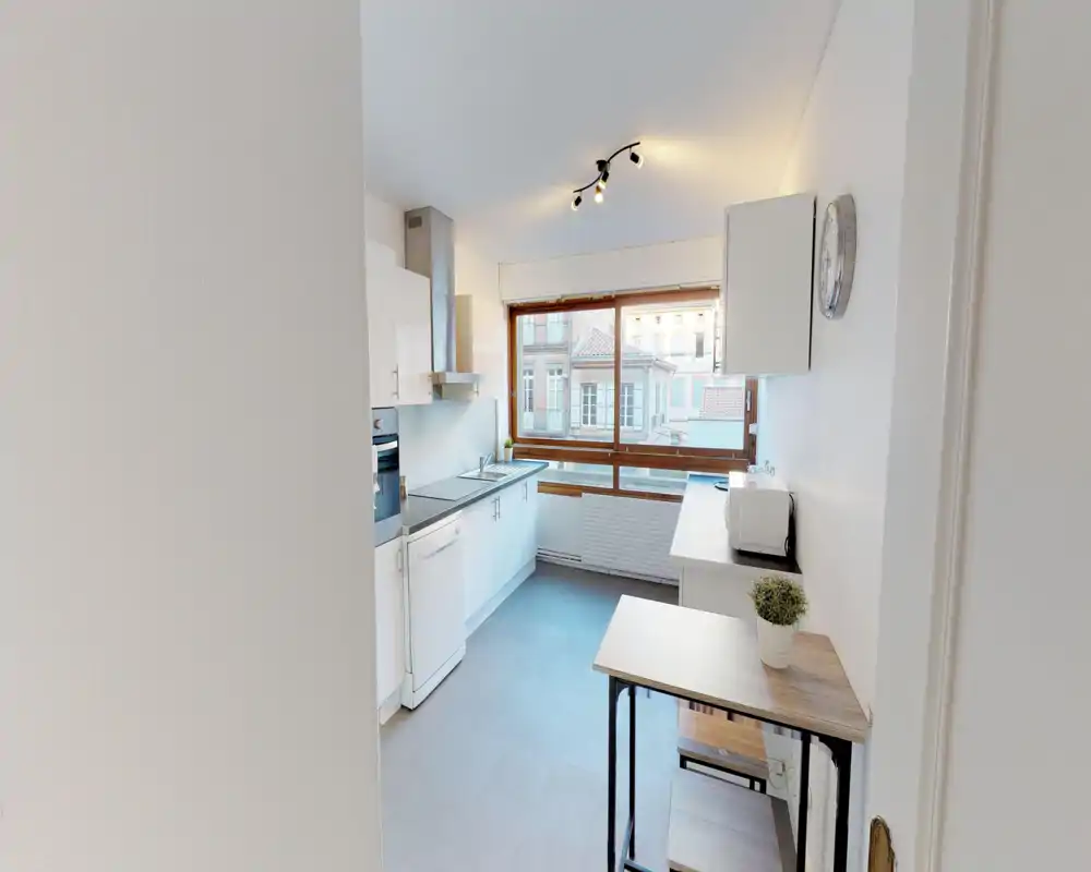 Bright modern kitchen with white cabinetry, built-in oven, dishwasher and a large window overlooking neighboring buildings. Clean minimalist surfaces, a small breakfast table and countertop appliances make this kitchen practical and welcoming.