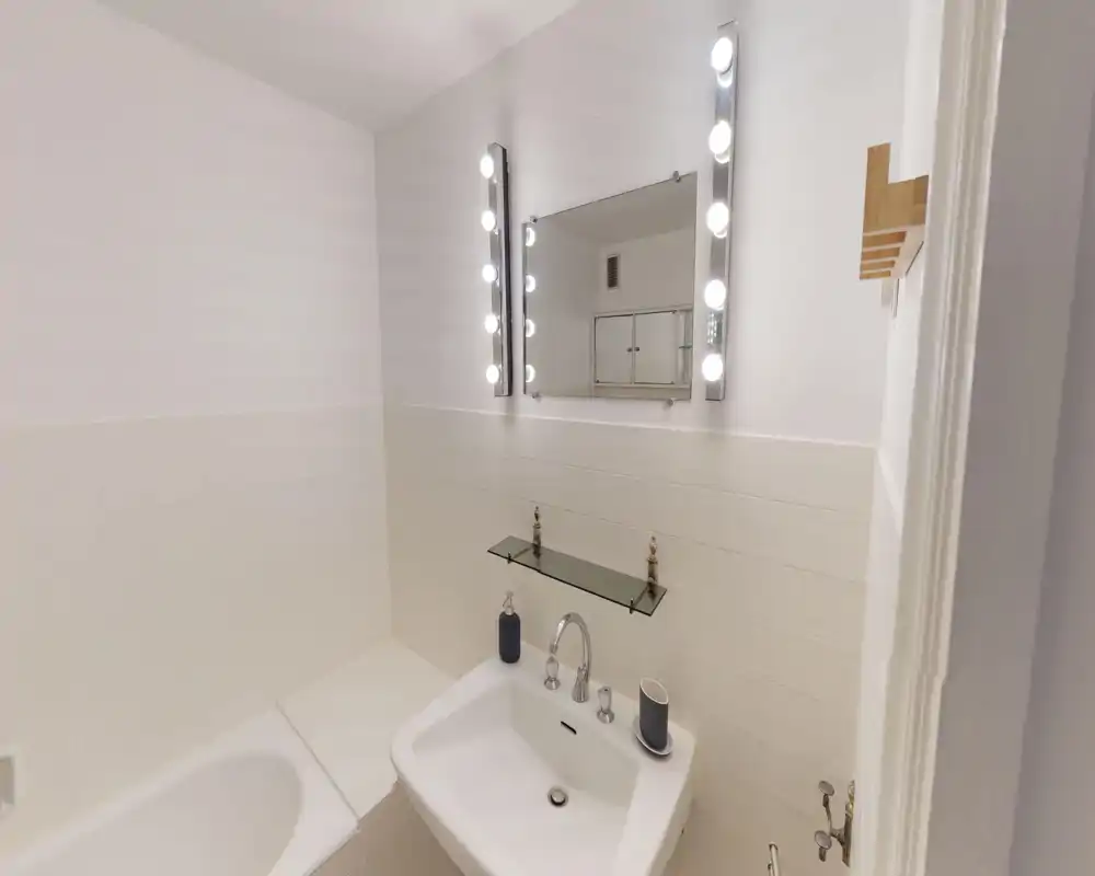 Clean, bright bathroom featuring a white sink with chrome fixtures, a vanity mirror flanked by lit vertical bulbs, and tiled walls above a built-in bathtub.