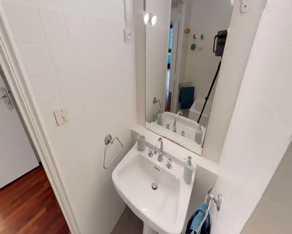 Compact, well-lit bathroom with a white pedestal sink, large mirror and chrome fixtures. Clean tiled walls and a towel rail visible next to the sink.