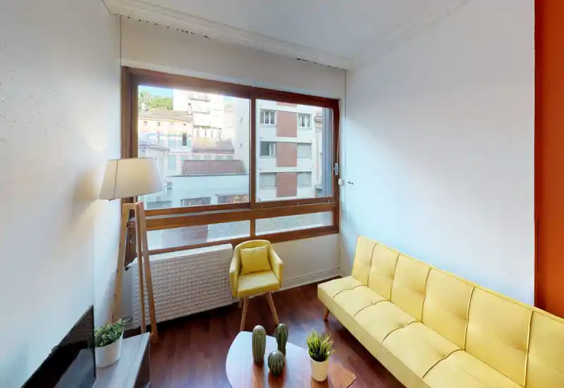Bright, modern living room with a large window, yellow sofa and matching armchair, wooden floor and decorative plants — a cozy, well-lit space ideal for relaxing.