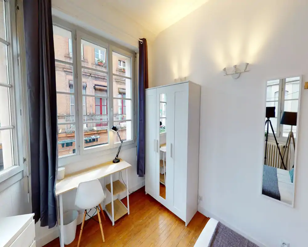 Coin chambre lumineux et bien aménagé avec parquet, grandes fenêtres, bureau et armoire blanche ; une partie du lit et le miroir sont visibles, ambiance chaleureuse et aérée.