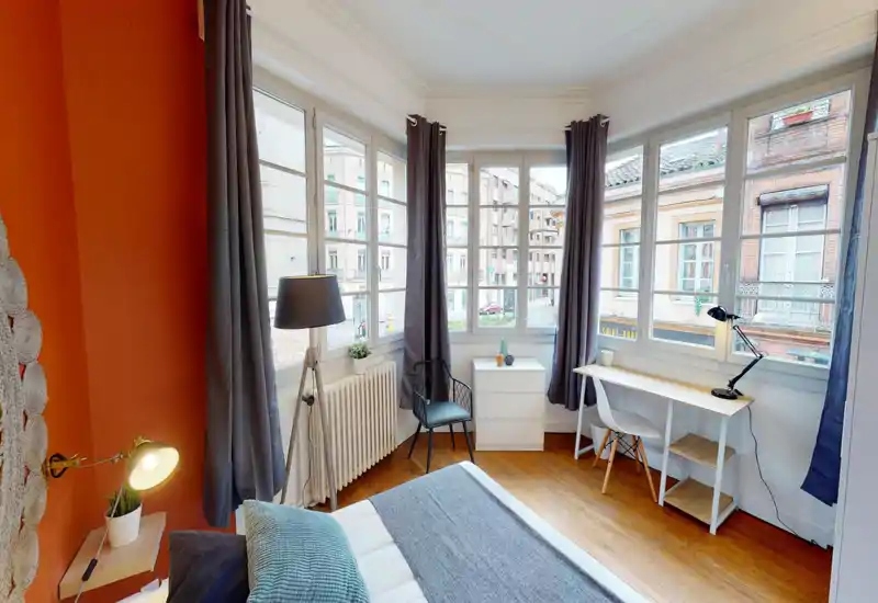 Chambre lumineuse et élégante avec une grande baie vitrée, des textiles chaleureux et un espace de travail près de la fenêtre. Le lit est clairement visible au premier plan et la pièce bénéficie d'une belle lumière naturelle.