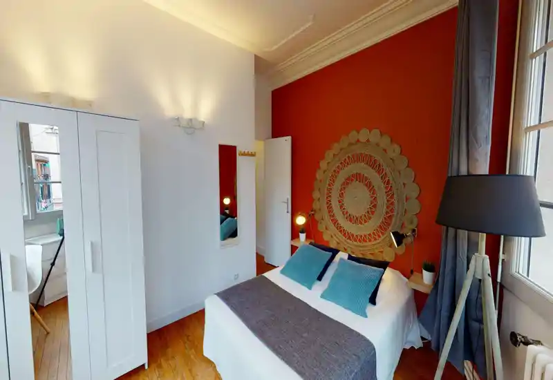 Chambre lumineuse et soignée avec un lit double en linge blanc et coussins bleus, une tête de lit tissée décorative sur un mur terracotta, lampes de chevet et un grand lampadaire près de la fenêtre.