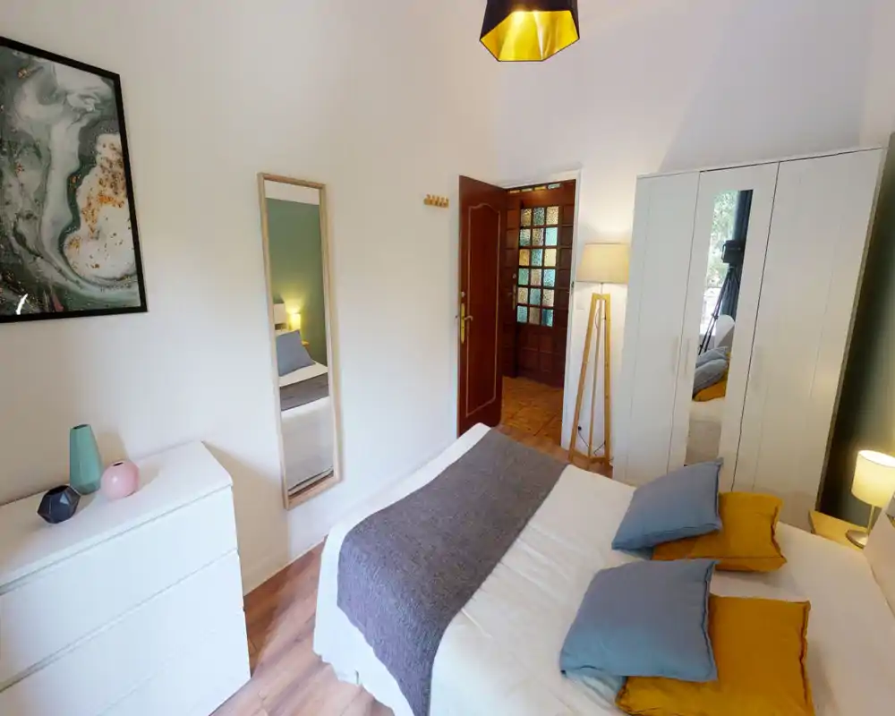 Bright, styled bedroom with a centrally framed double bed, decorative pillows and a warm floor lamp; large wardrobe and dresser provide practical storage.