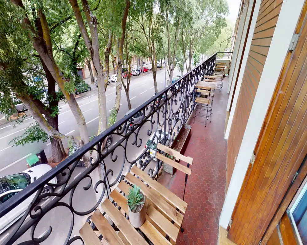 Sunny narrow balcony with a decorative wrought-iron railing, wooden bistro tables and chairs, small potted plants, and a clear view over a tree-lined street.
