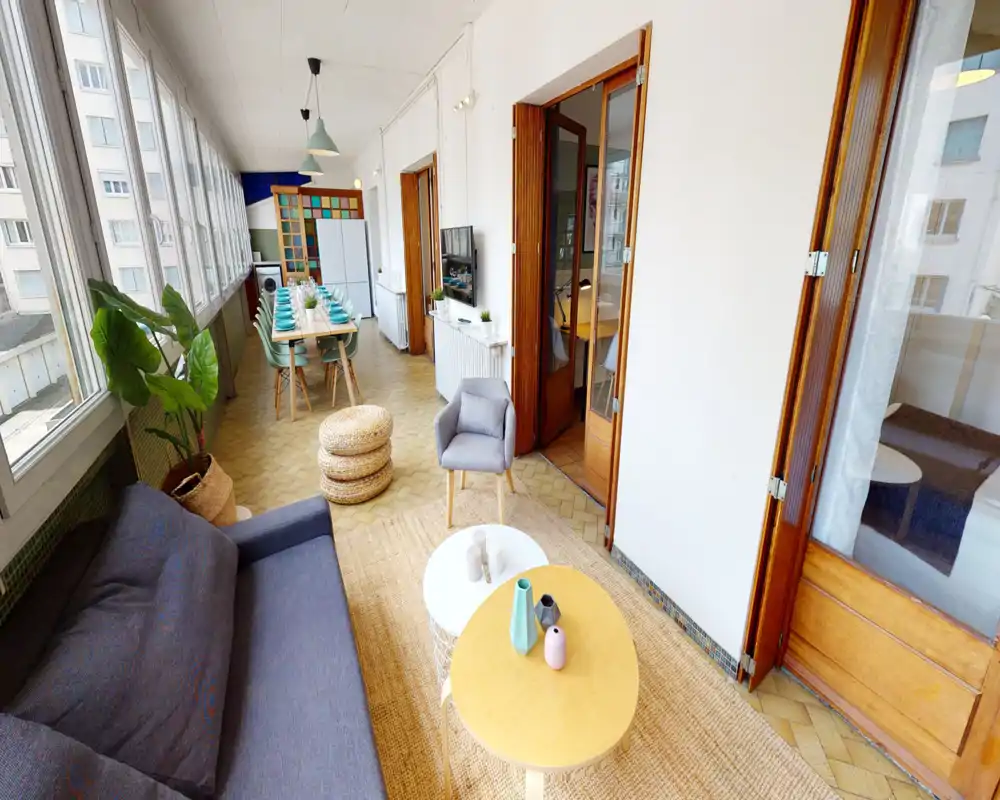 Bright, stylish living area with a comfortable sofa, small coffee tables and natural light from large windows; a dining area is visible further down the enclosed balcony space.