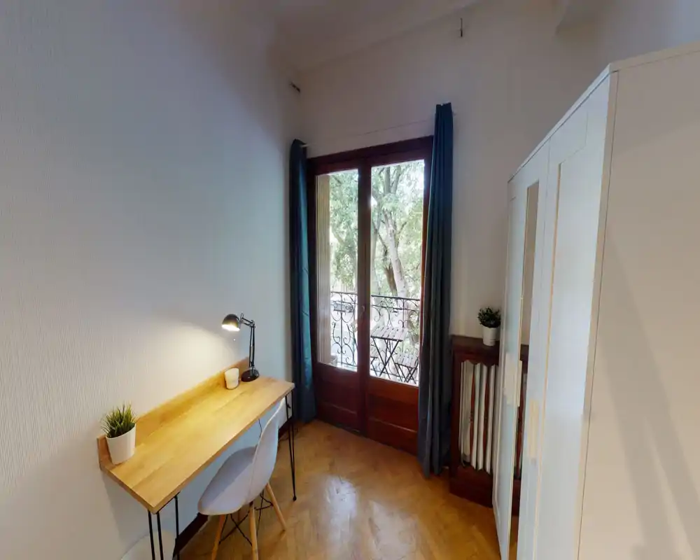 Bright, tidy bedroom corner with a wooden desk, chair and wardrobe beside a balcony door that opens onto a small outdoor seating area. The image highlights a calm workspace and large windows that bring natural light.