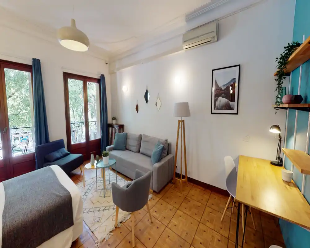 Bright, stylish bedroom with a comfortable double bed in the foreground, a seating area with sofa and armchair, and a work desk by the wall. Large windows provide abundant natural light and a leafy view.