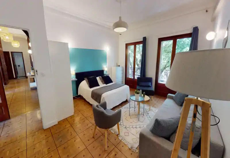 Bright, stylish bedroom with a double bed, teal accent wall and seating area; large windows bring in natural light and a cozy rug anchors the space.