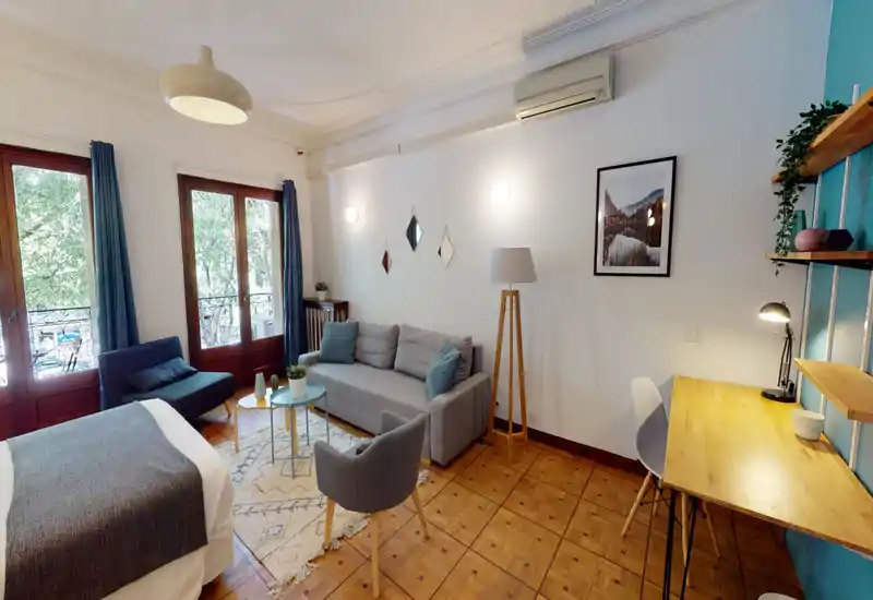 Bright, stylish bedroom with a comfortable double bed in the foreground, a seating area with sofa and armchair, and a work desk by the wall. Large windows provide abundant natural light and a leafy view.