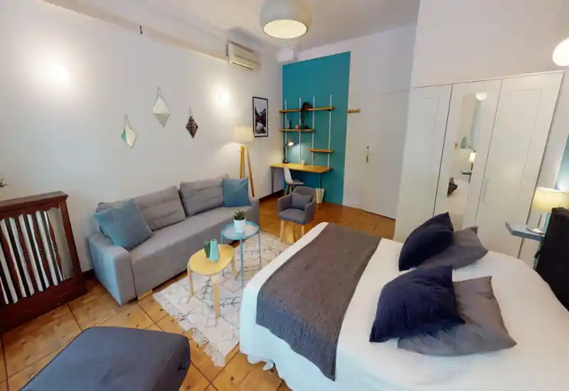 Bright, stylish bedroom with a large bed in the foreground, cozy textiles and coordinated decor. The room includes a sofa and a small desk area, creating a comfortable studio-like layout.