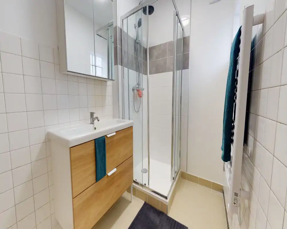 Bright, modern bathroom with a corner glass shower, wall-mounted vanity with wooden drawers and a mirrored cabinet above. Clean tiled walls and a heated towel rail with a teal towel complete the space.