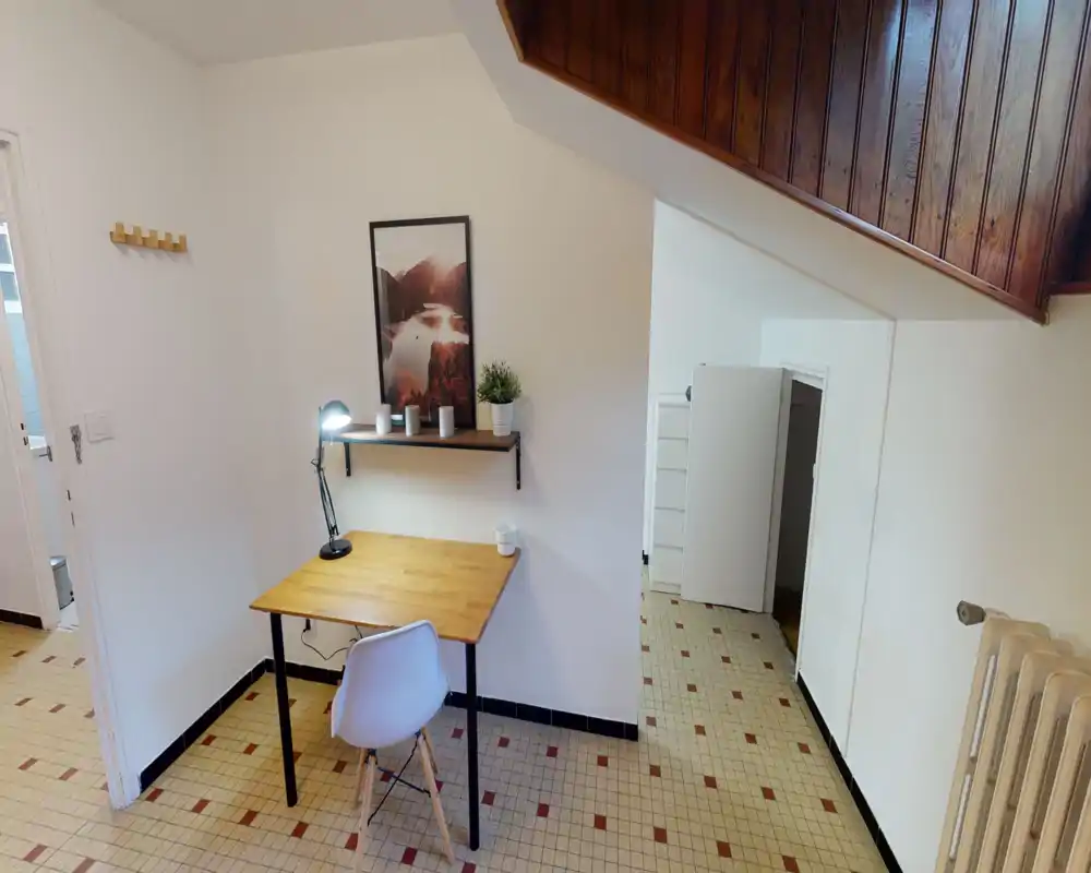 Compact bright workspace with a wooden desk, modern chair and a shelf-mounted lamp; minimal décor includes a framed print and a small plant.