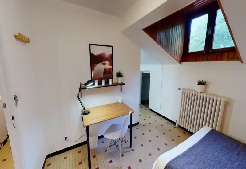 Compact, bright bedroom corner with a single bed, a wooden desk and skylight. The space features simple modern decor, framed art, a desk lamp and potted plants, ideal for a comfortable stay or student rental.