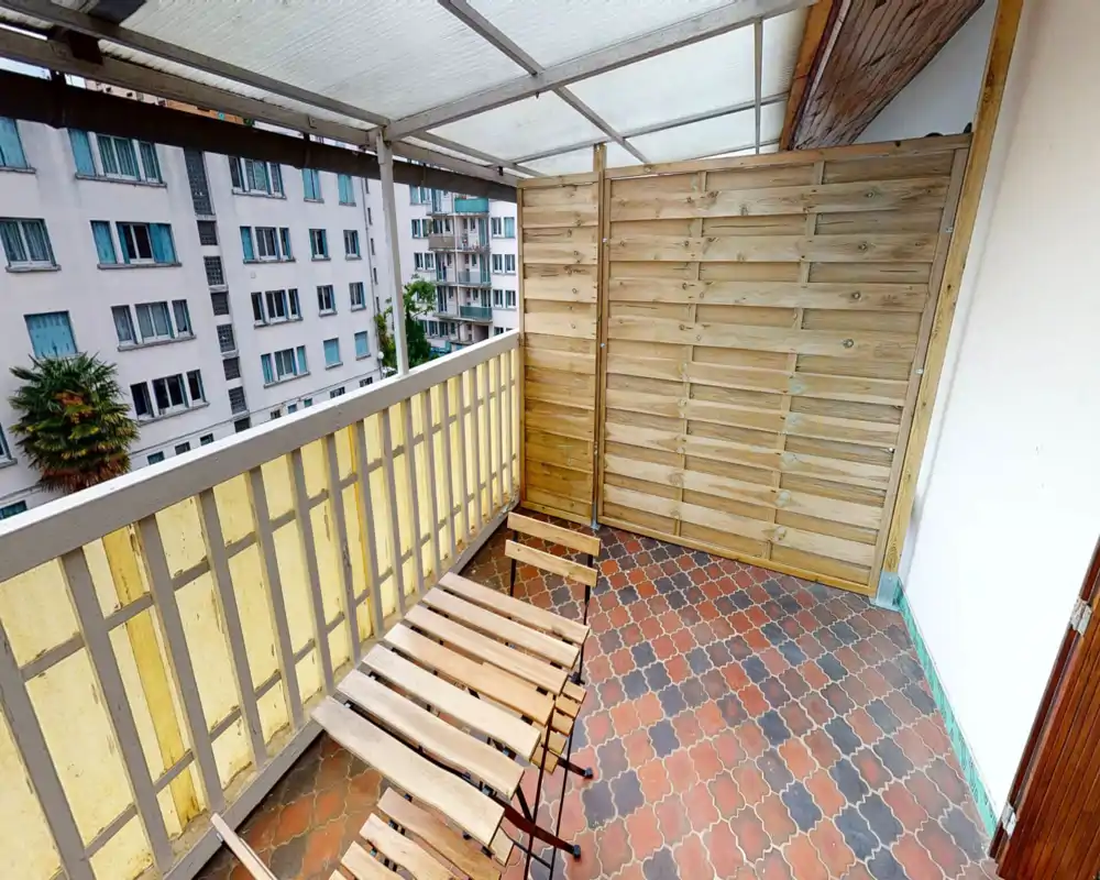 Covered balcony with patterned floor tiles, wooden privacy screen and a small foldable wooden table and chairs — bright, sheltered outdoor space overlooking neighboring buildings.