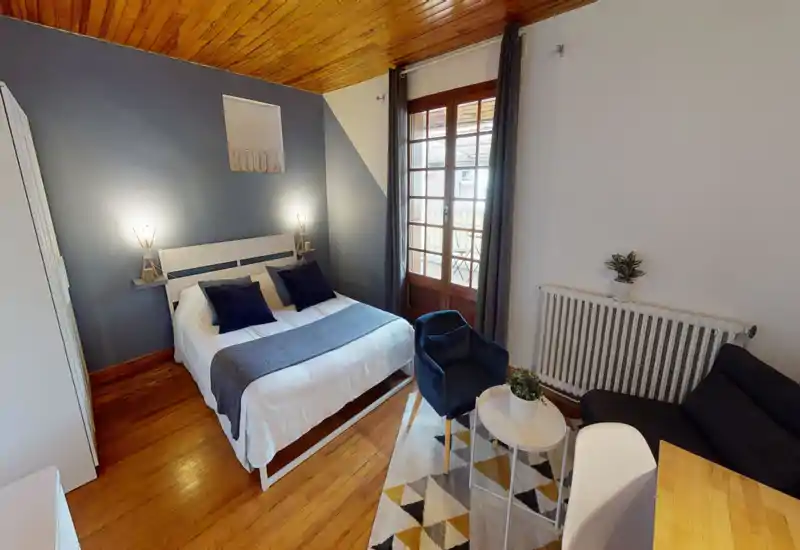 Bright, well-styled bedroom with a double bed, bedside lamps, wooden ceiling and a small seating area by the window. Neutral and blue tones create a calm, cozy atmosphere ideal for rental photos.