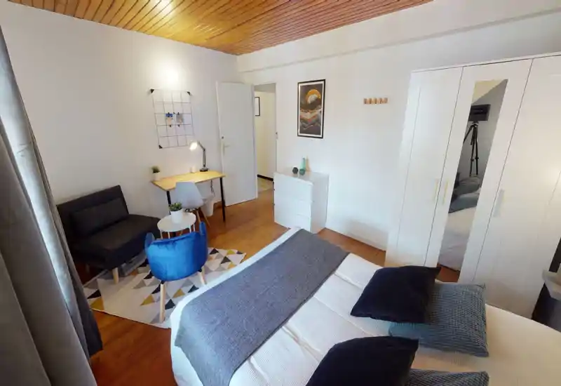 Bright, well-staged double bedroom with a comfortable bed in the foreground, a seating corner with a blue accent chair and a small desk by the wall — ideal for short-term rentals or students.