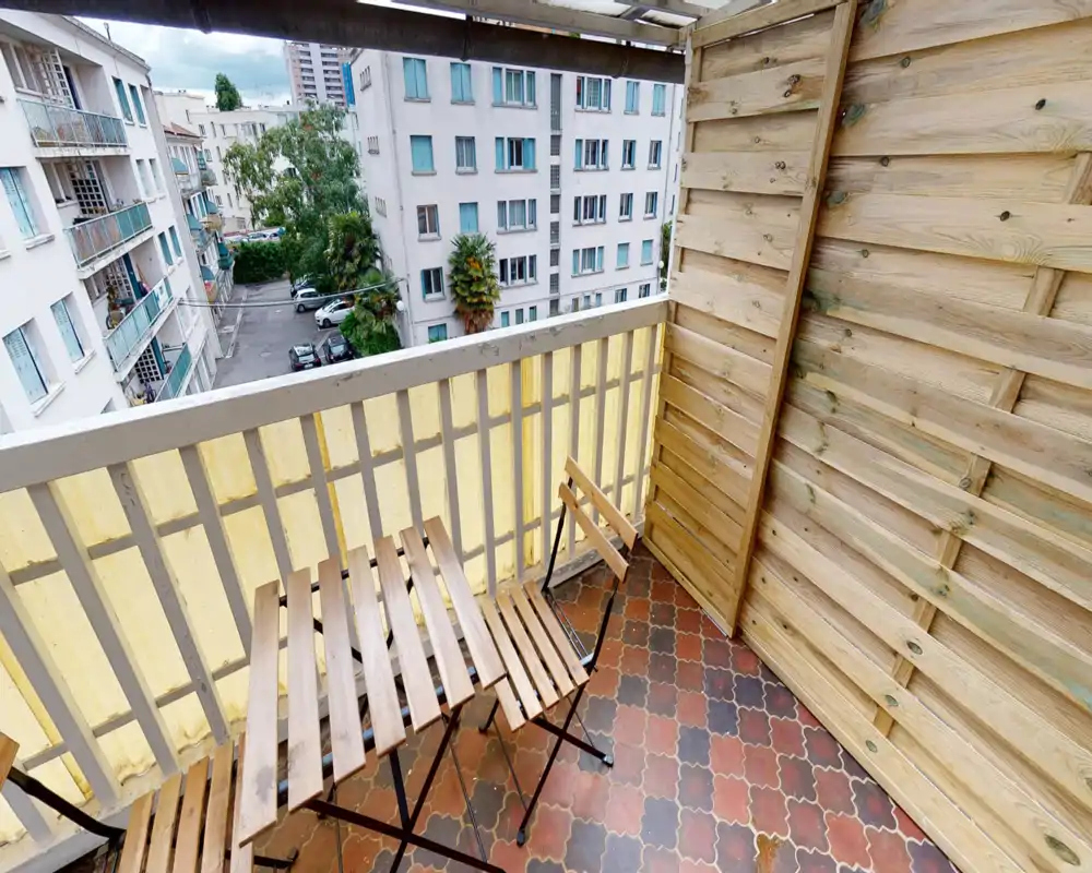 Piccolo balcone soleggiato con sedie e tavolo in legno a doghe, pavimento piastrellato e pannelli di legno per la privacy; vista sui palazzi e sul cortile.