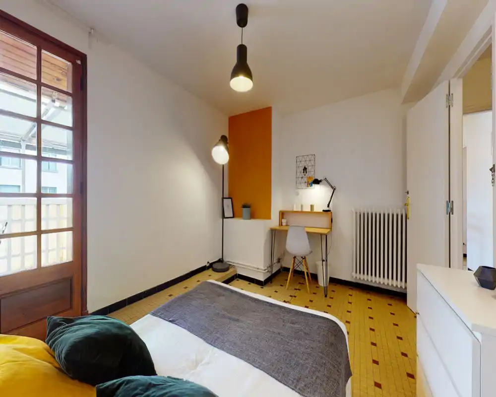Camera da letto luminosa e accogliente con letto matrimoniale, cuscini decorativi e una piccola scrivania lungo la parete. Ampia porta finestra che dona luce naturale e collegamento al balcone.