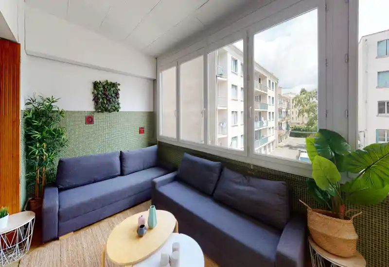 Bright enclosed balcony styled as a cozy living area with two modern grey sofas, low nesting coffee tables and abundant indoor plants. Large windows overlook neighboring buildings and fill the space with natural light.