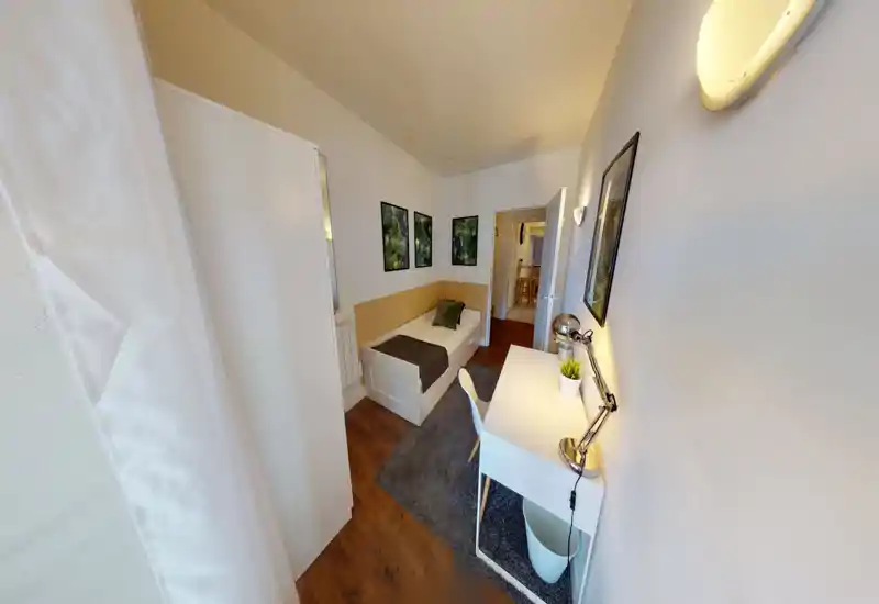 Compact, well-styled single bedroom with a neatly made bed, desk with lamp and a soft rug — bright and ready for a guest.