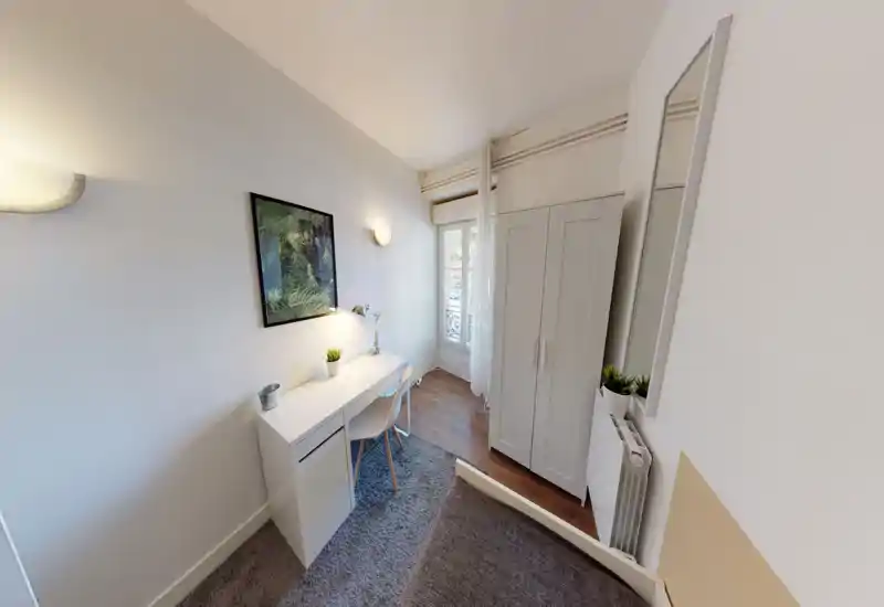 Compact, bright bedroom with a small desk by the window, wardrobe and a partially visible bed. Clean, modern styling with natural light and simple decorative plants.