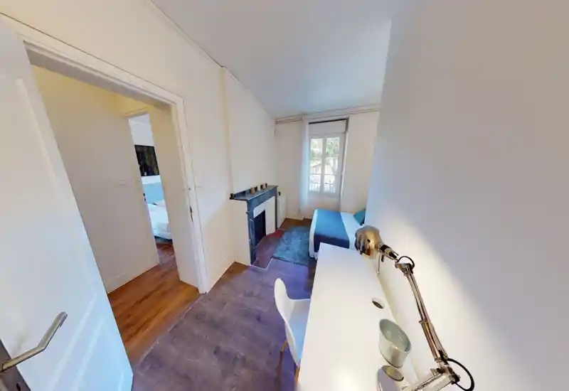 Bright, simply styled bedroom with a double bed, desk and window. The room features a small decorative mantle and wooden floors, offering a clean and practical space for rest and work.