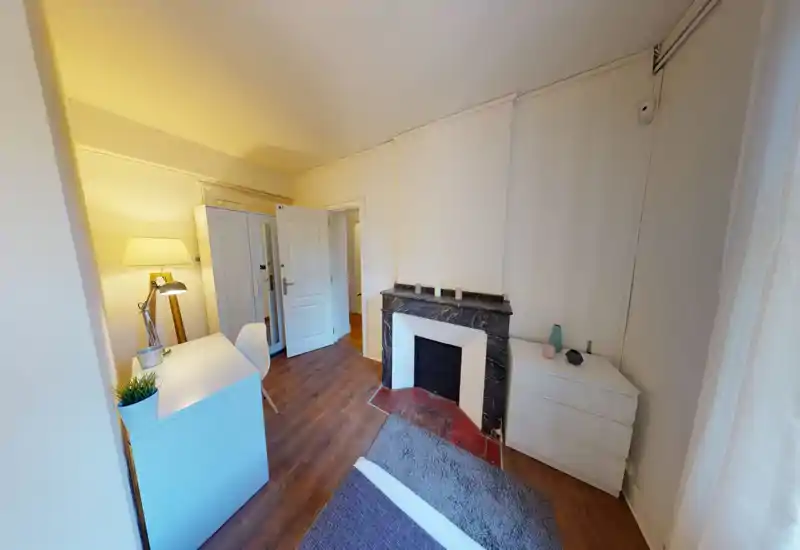 Cozy bedroom with a visible bed edge and soft rug, a desk with lamp, wardrobe and a decorative fireplace — warm, well-lit and functional for guests or long stays.