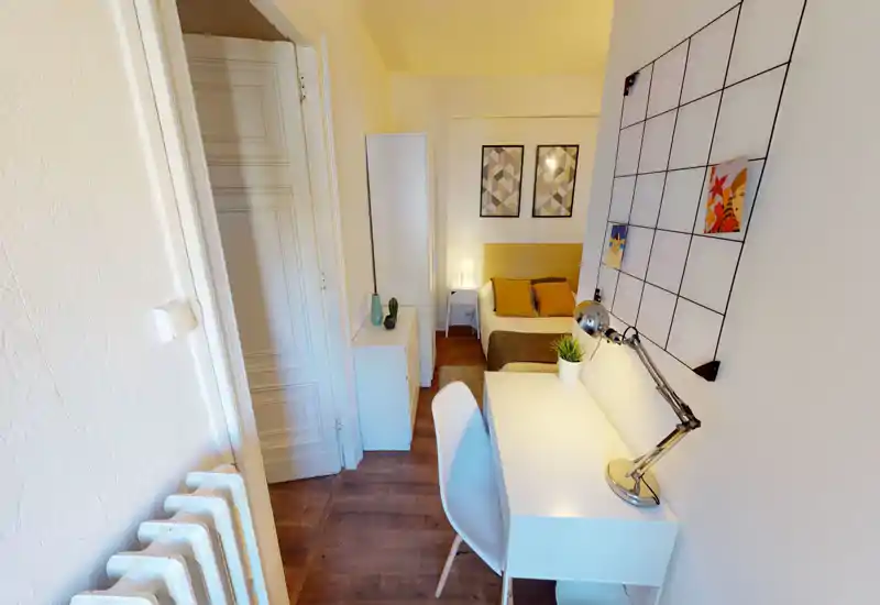 A compact, well-styled bedroom with a double bed, bedside table and a neat work desk by the wall; warm lighting and coordinated cushions create a cozy atmosphere.