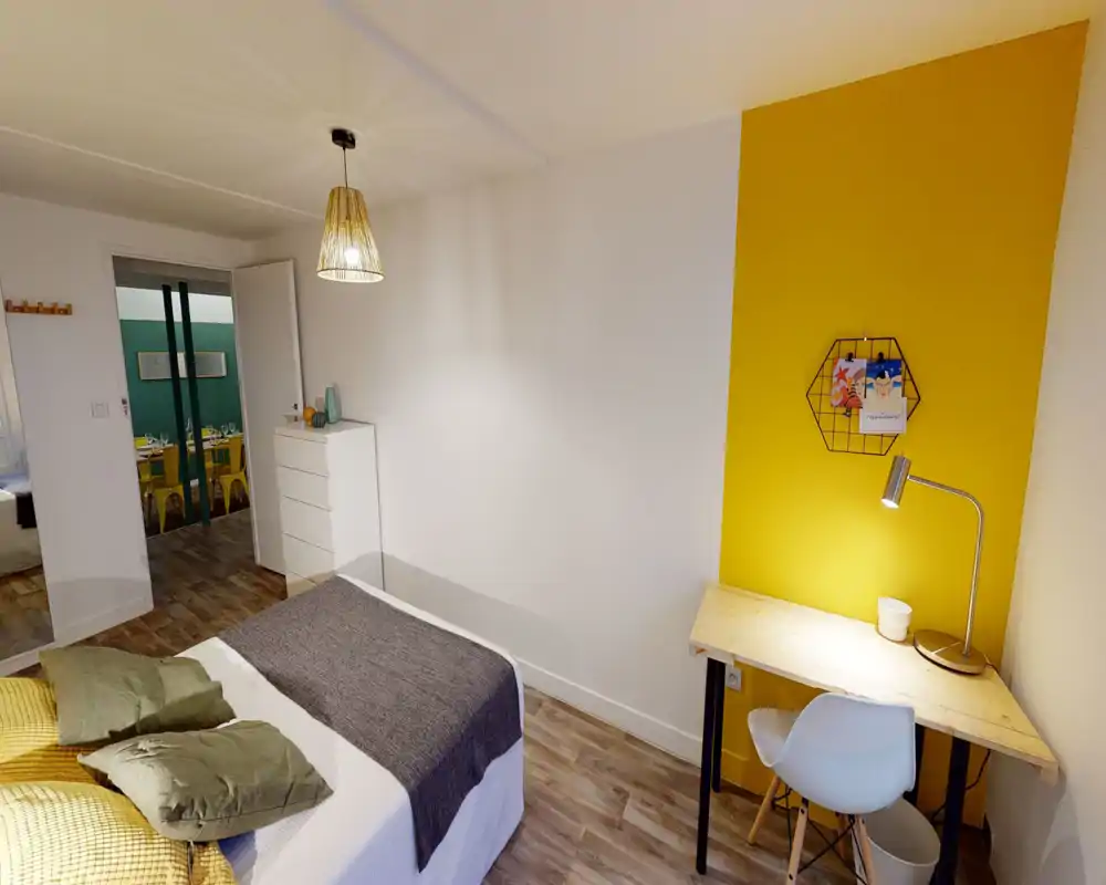 Bright, modern bedroom with a comfortable double bed, textured throws and cushions, a wooden desk with a lamp against a warm yellow accent wall, and a chest of drawers. Clean lines and cozy textiles make it ideal for short-term rentals.
