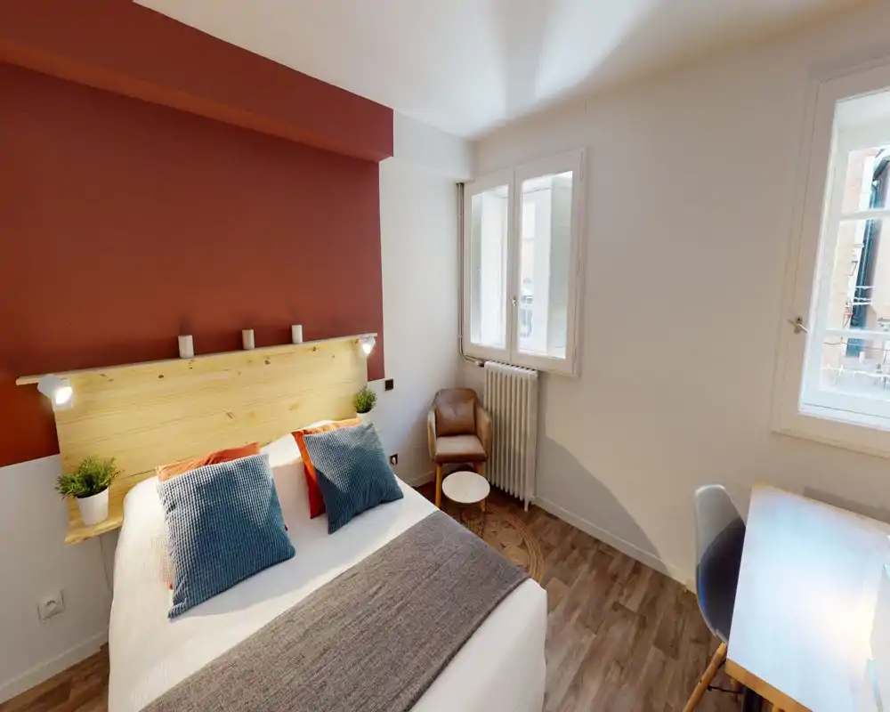 Bright, well-styled bedroom with a double bed, textured pillows, a warm accent wall and natural light from two windows; includes a small armchair and desk for a comfortable stay.