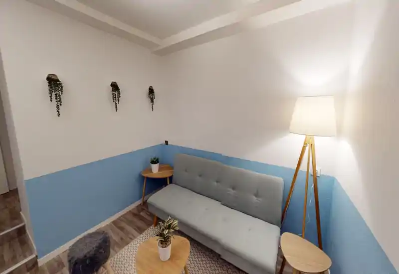 Petit salon soigné avec canapé gris clair, tables en bois, plantes décoratives et lampe sur pied chaleureuse — cosy et bien mis en scène pour la location courte durée.