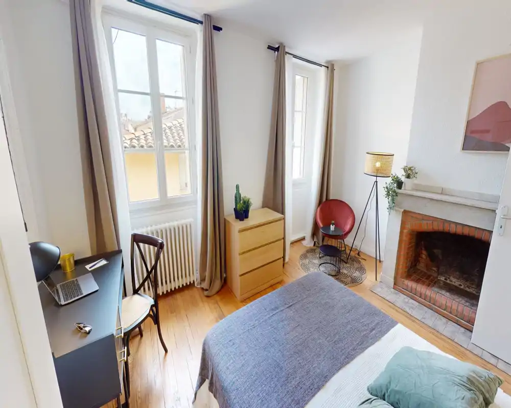 Dormitorio luminoso y bien decorado con una cama grande en primer plano, dos ventanas altas que aportan luz natural, suelos de madera y una chimenea decorativa. Un pequeño escritorio junto a la ventana y un rincón de lectura con lámpara y butaca completan el espacio acogedor.
