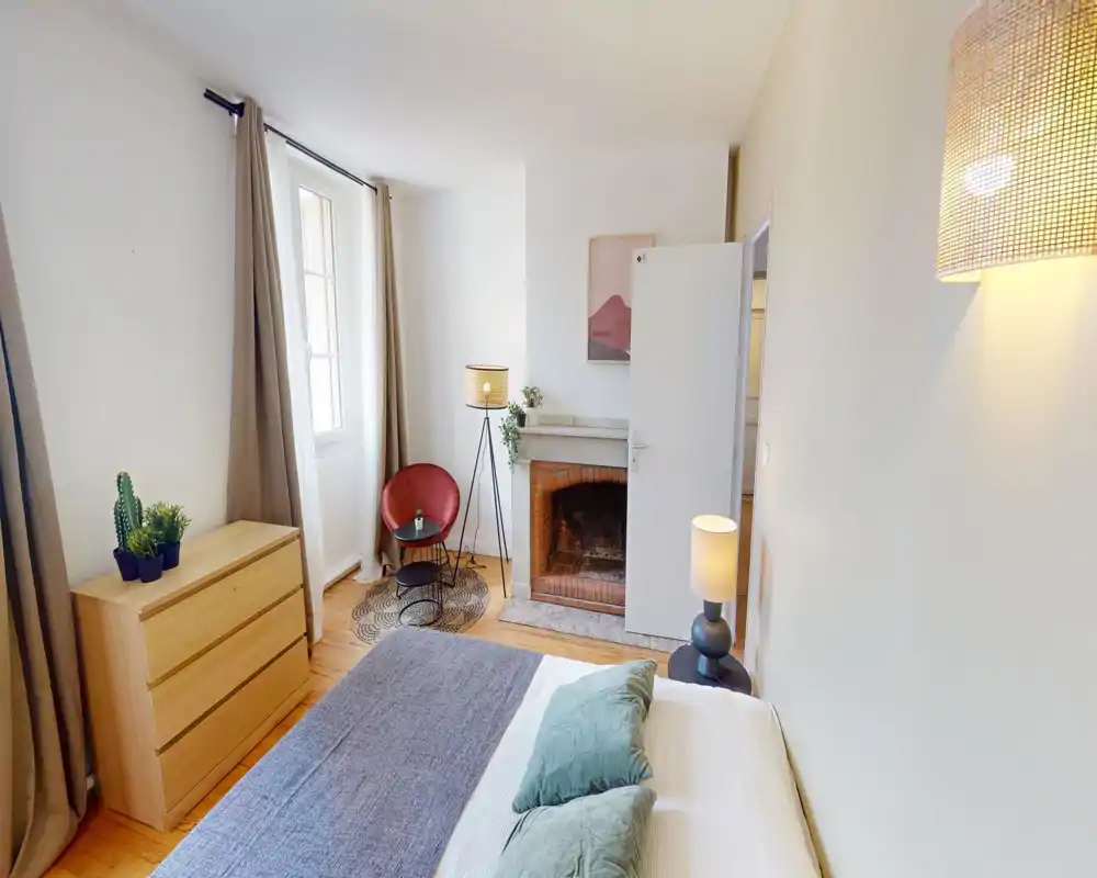 Dormitorio luminoso y con estilo, con una cama grande vestida en tonos neutros, cojines verdes, cómoda de madera y un rincón acogedor con lámpara y butaca junto a una pequeña chimenea.