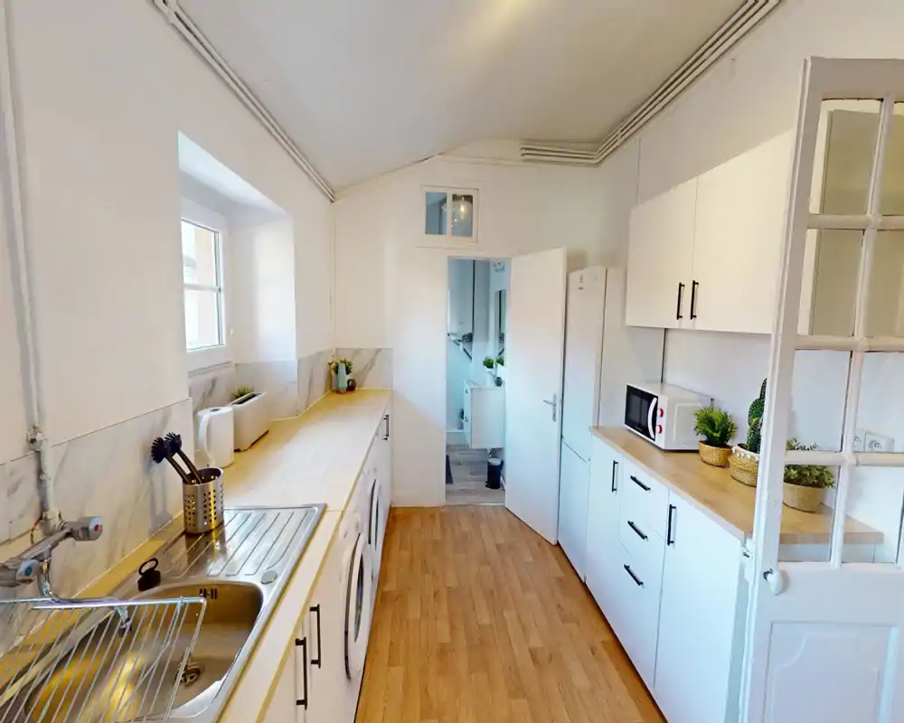 Cucina luminosa e stretta con pensili bianchi, piani in legno, elettrodomestici integrati e lavello in acciaio; ordinata e ben allestita per foto di affitto.
