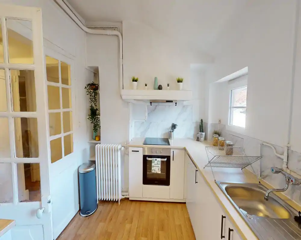 Cucina luminosa e compatta con mobili bianchi, forno e piano cottura integrati, piani in legno e lavello in acciaio. Spazio pulito e ben illuminato con piccole piante e contenitori sul piano.