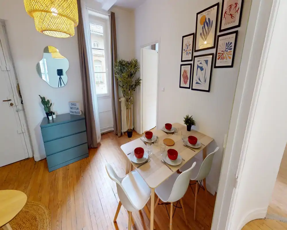 Zona pranzo luminosa e arredata con tavolo in legno per quattro persone, sedie bianche moderne e quadri alle pareti; la luce naturale e il pavimento in legno creano un ambiente accogliente.