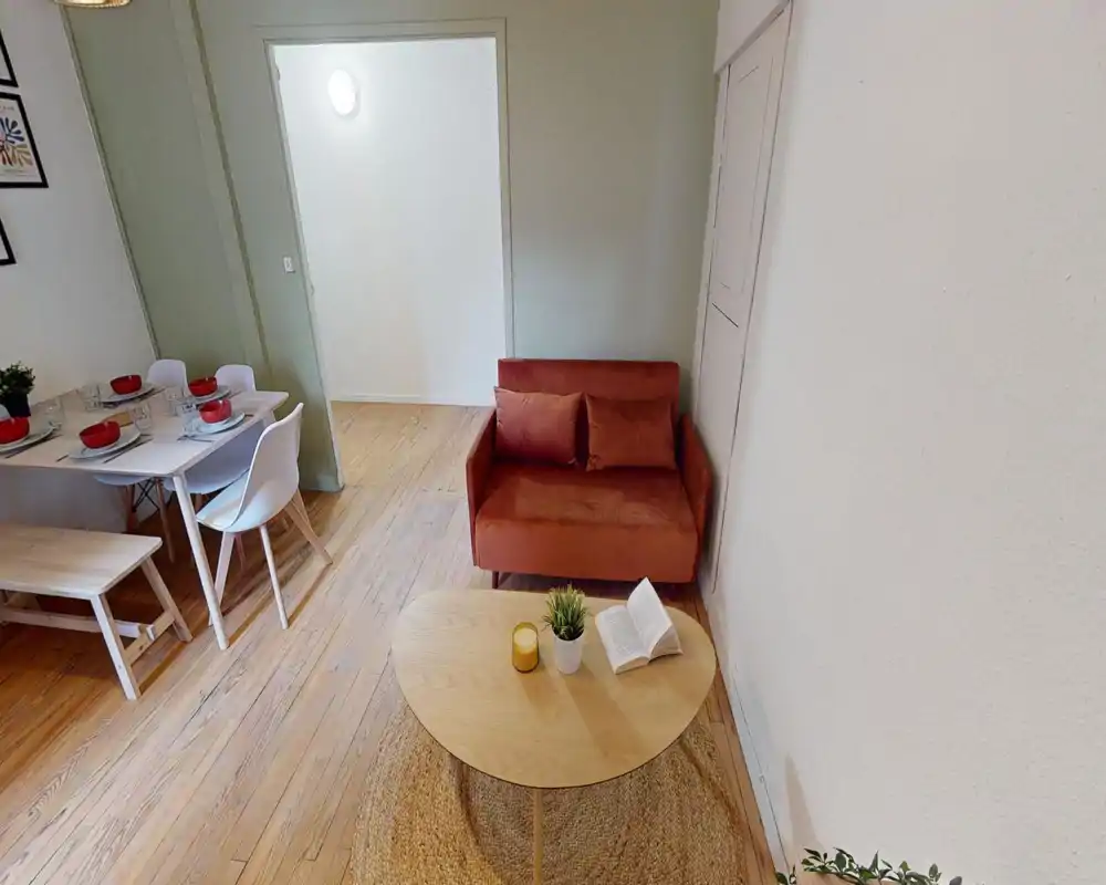 Zona giorno accogliente con un divanetto color ruggine, un tavolino in legno su un tappeto in juta e un angolo pranzo visibile a lato. Pavimenti in legno caldo e dettagli decorativi rendono l'ambiente invitante.